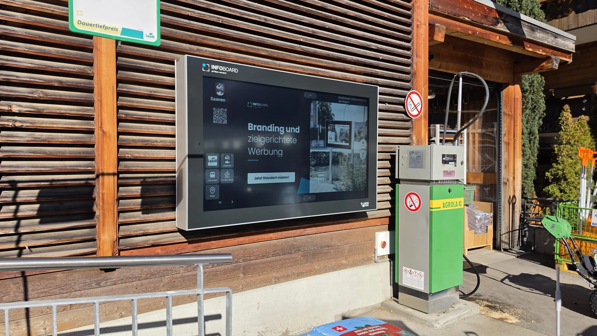 Pantalla exterior Infoboard de 75 pulgadas en la fachada de madera de la Landi en la estación de Gstaad, con botones para Saanen, horarios y mapa