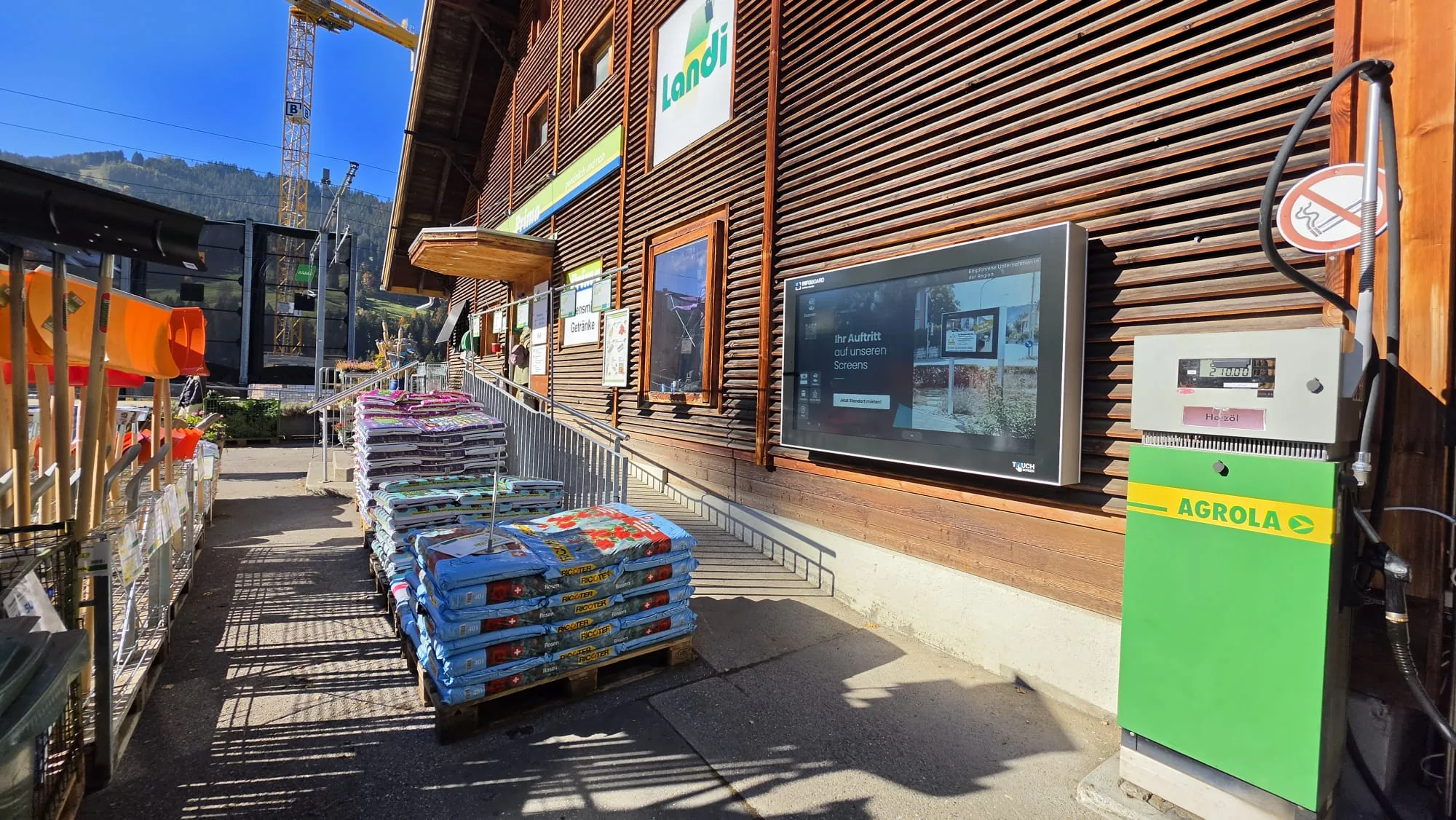 La pantalla exterior de la Landi junto a la estación de Gstaad vista de lejos, con la explanada, la escalera y las montañas al fondo