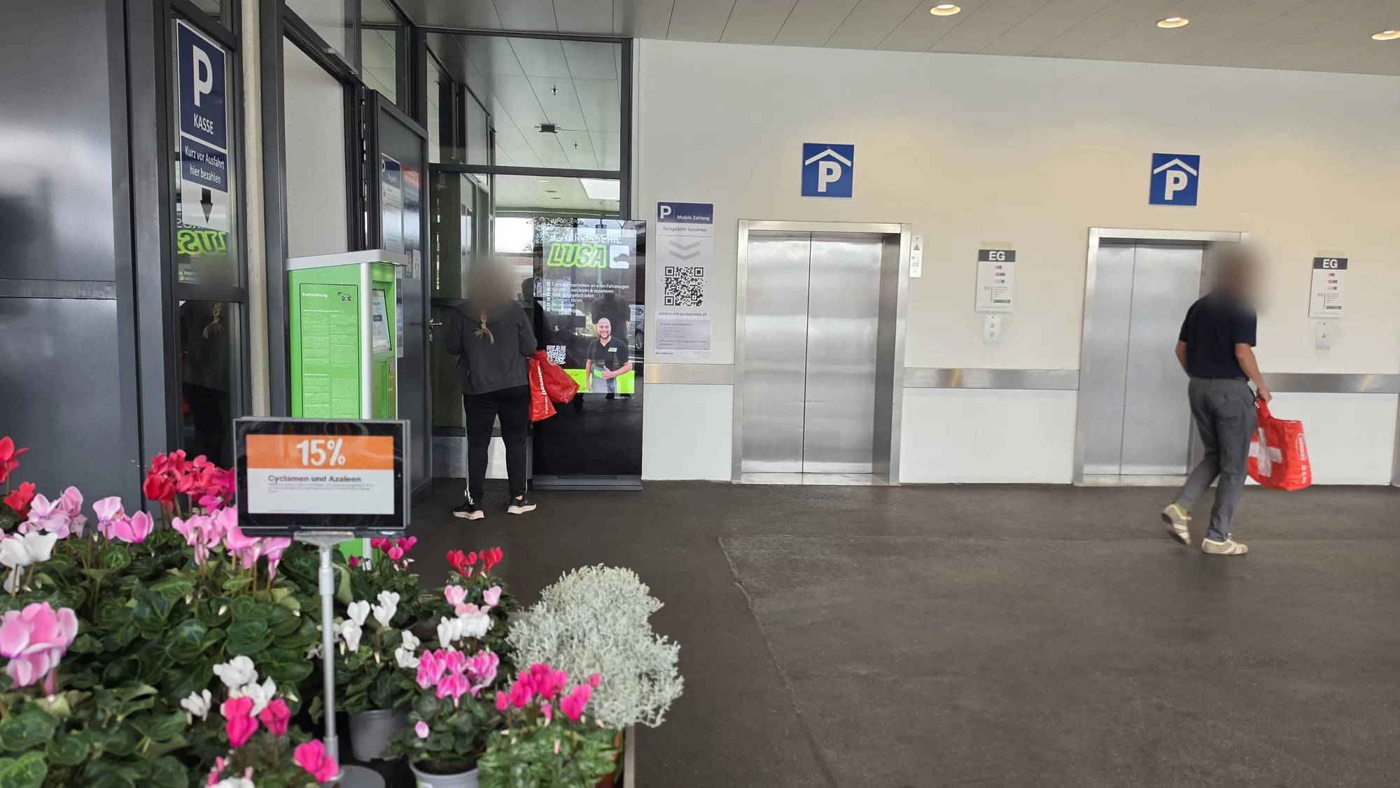Ingresso della Migros di Grenchen con schermo pubblicitario e clienti diretti al parcheggio