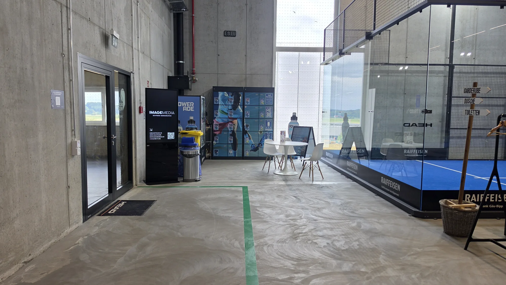 Ingresso del centro padel Padelwerk con totem di digital signage accanto alla porta e campo con pareti in vetro