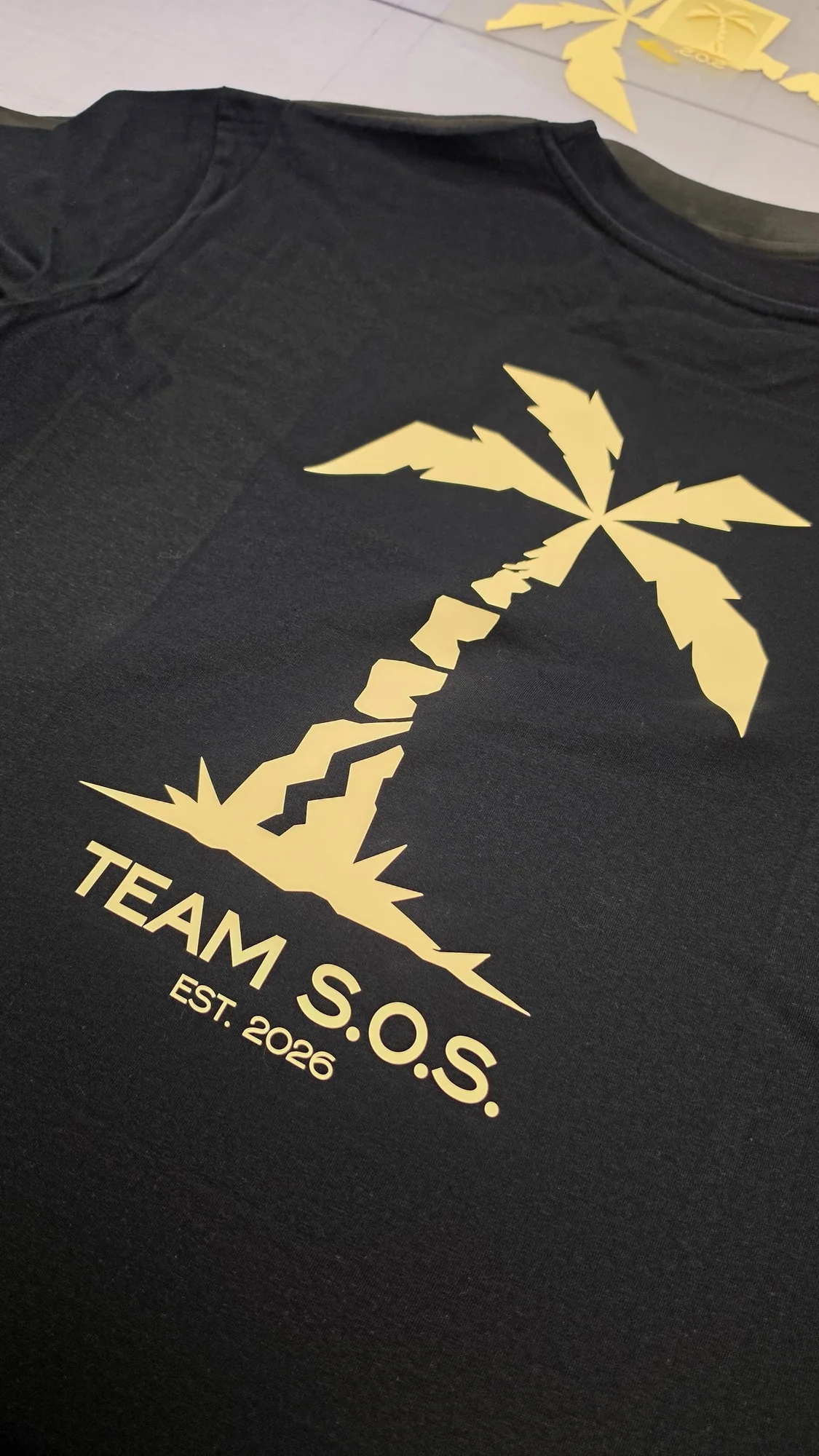 Camiseta negra con logo «Team S.O.S. Est. 2026» y palmera en vinilo flex amarillo